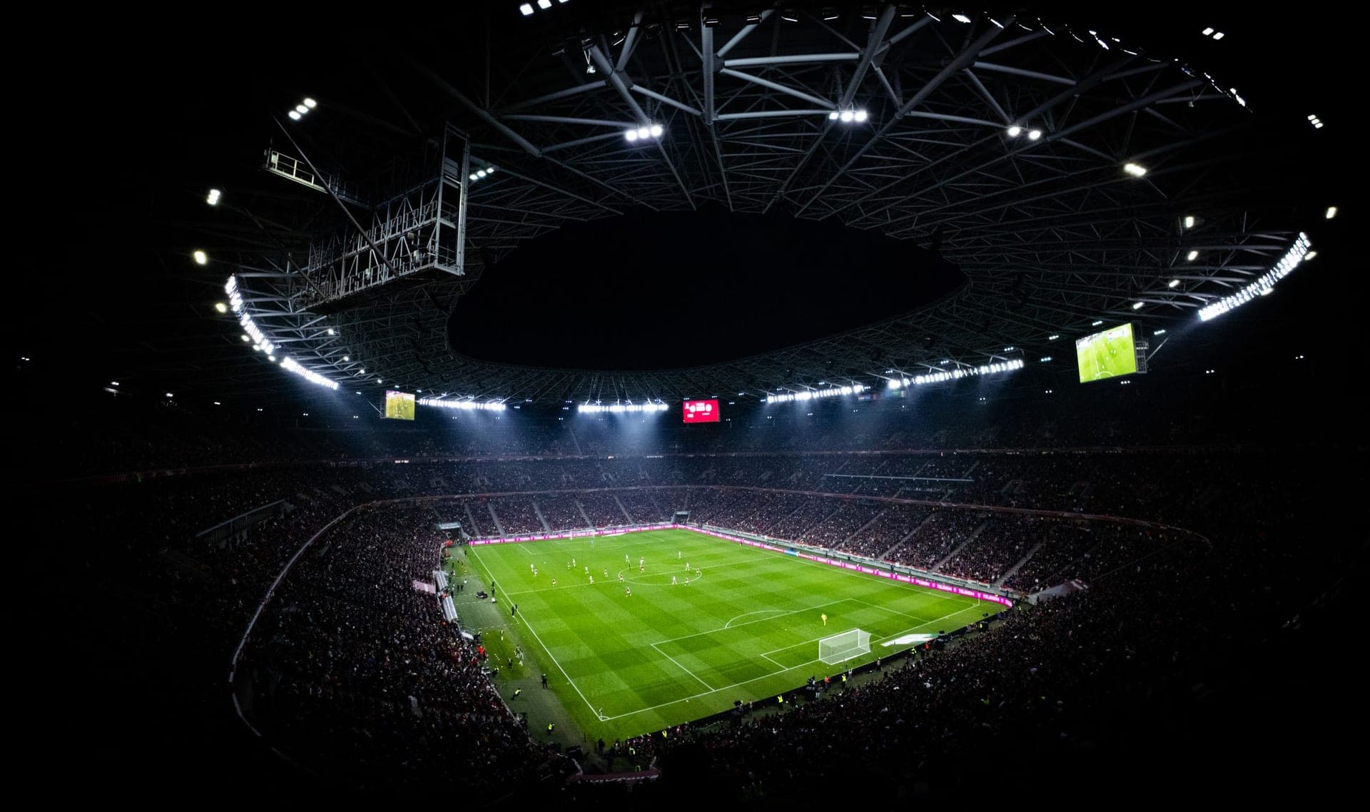 A packed soccer stadium under floodlights at night, the pitch glowing green in the dark
