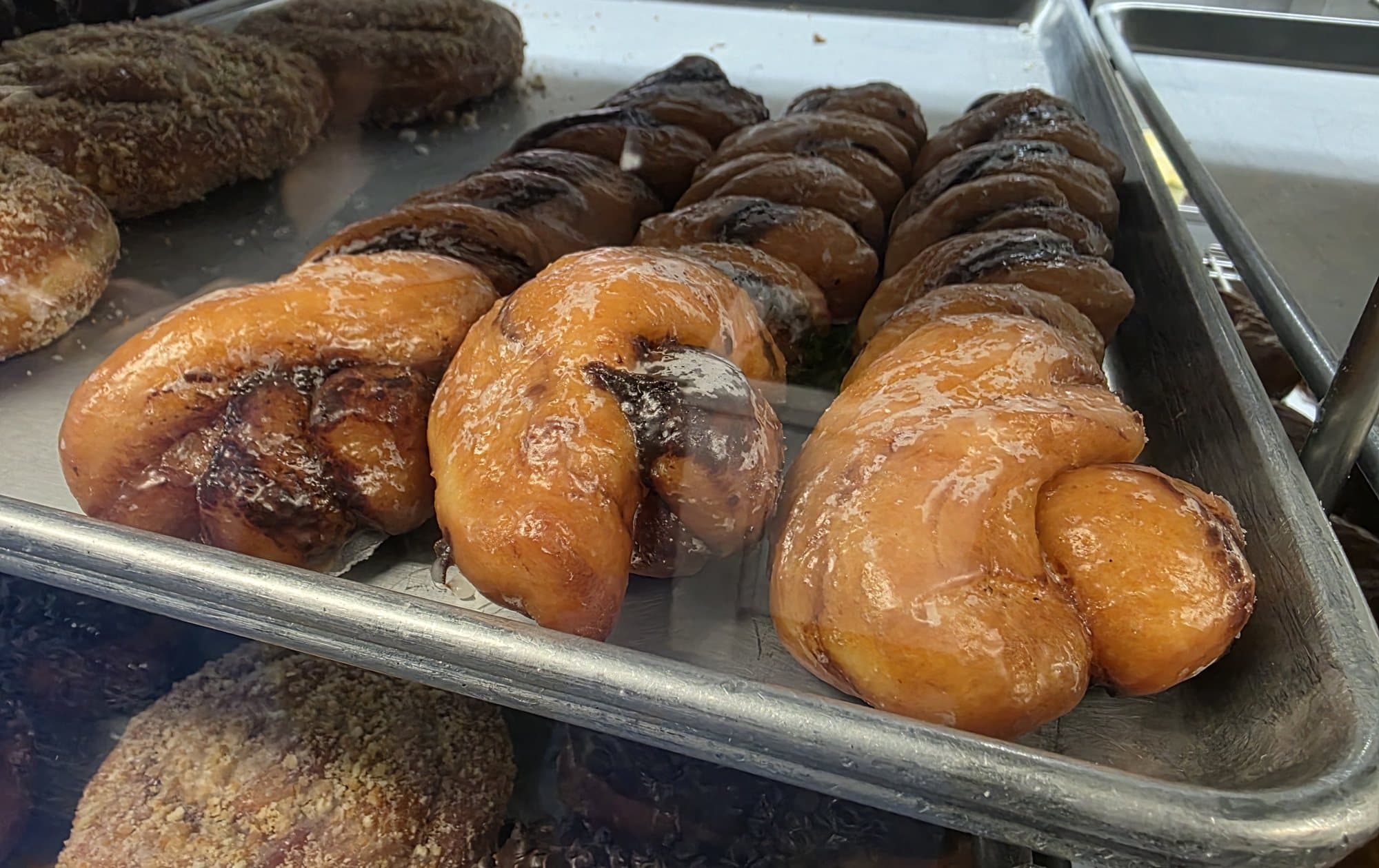 Glazed twist donuts and crumb-topped donuts on a metal tray in the case at Rolling Pin Donuts in Camarillo