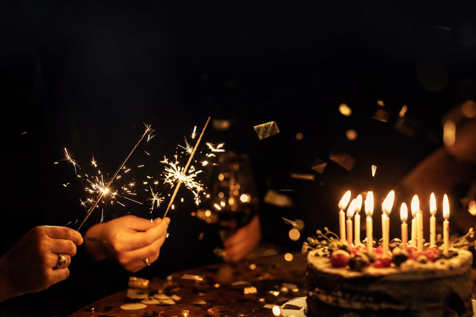 Sparklers, lit birthday candles on a cake, and gold confetti against a deep warm background