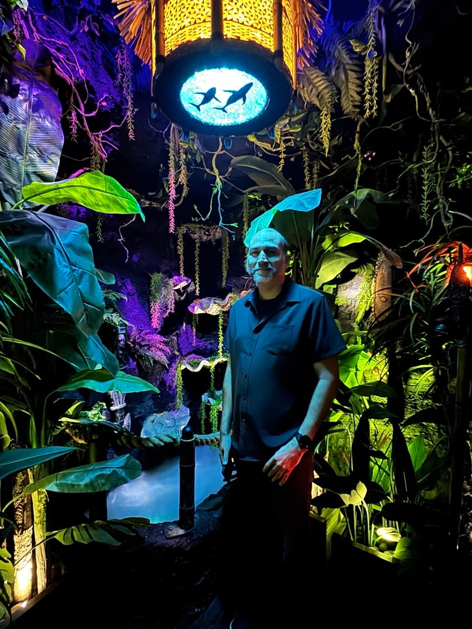 Jason Walters standing under the aquarium archway at House of Bamboo, with cyan-lit orca silhouettes glowing overhead and lush tropical plants framing the space
