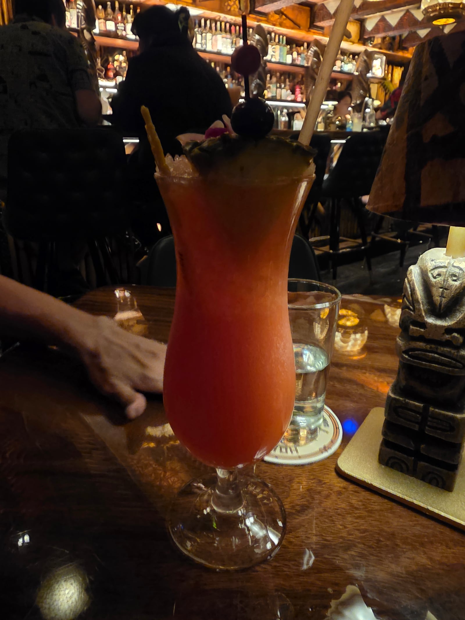 A Hurricane cocktail in a tall hurricane glass at House of Bamboo in Camarillo, with a pineapple wedge and cherry garnish, a bamboo stir straw, and the lit bar shelves in the background