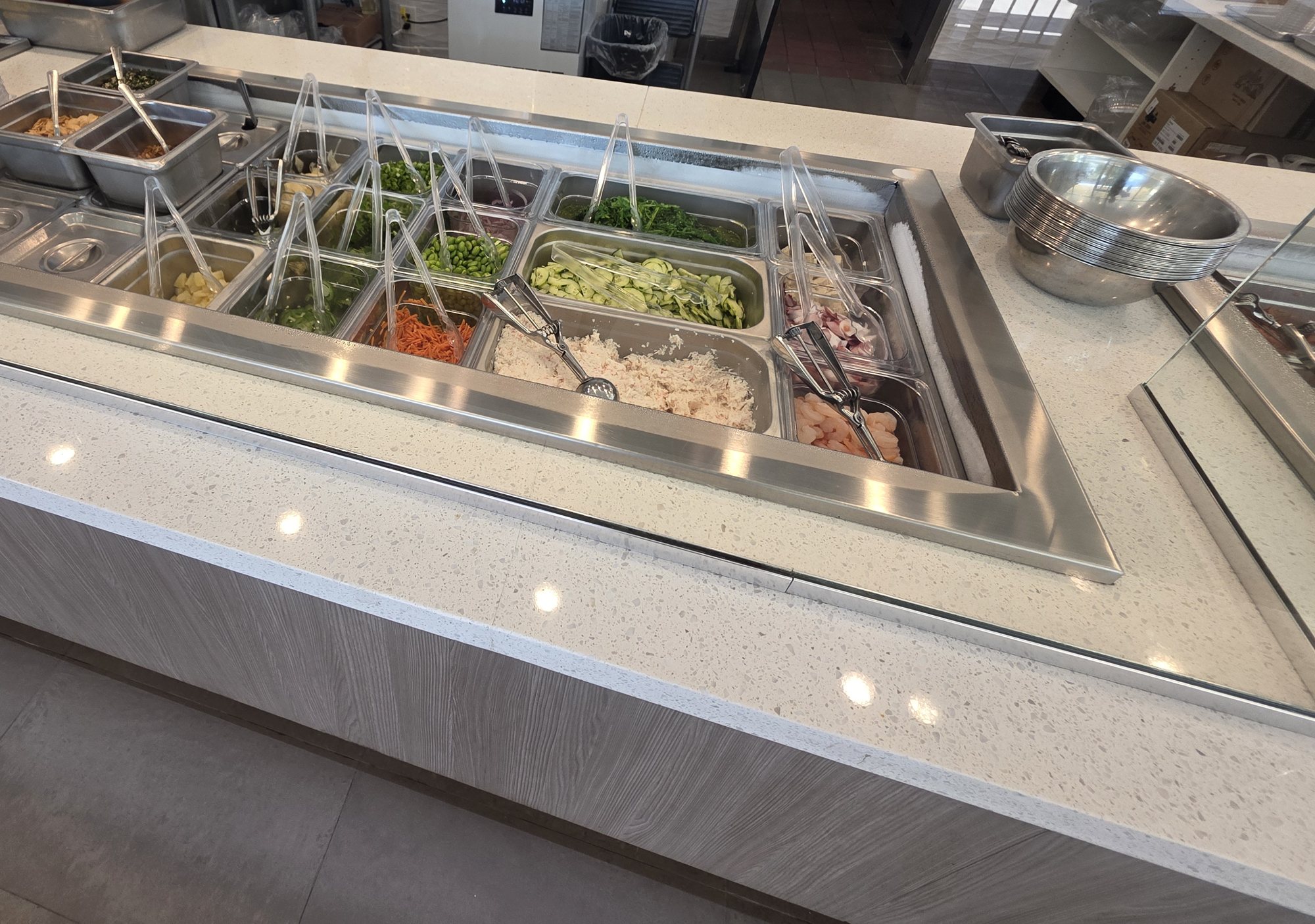 The sides and toppings bar at Poke & K-Roll, with edamame, cucumber, shredded carrot, crabmeat, onion and more in steel pans