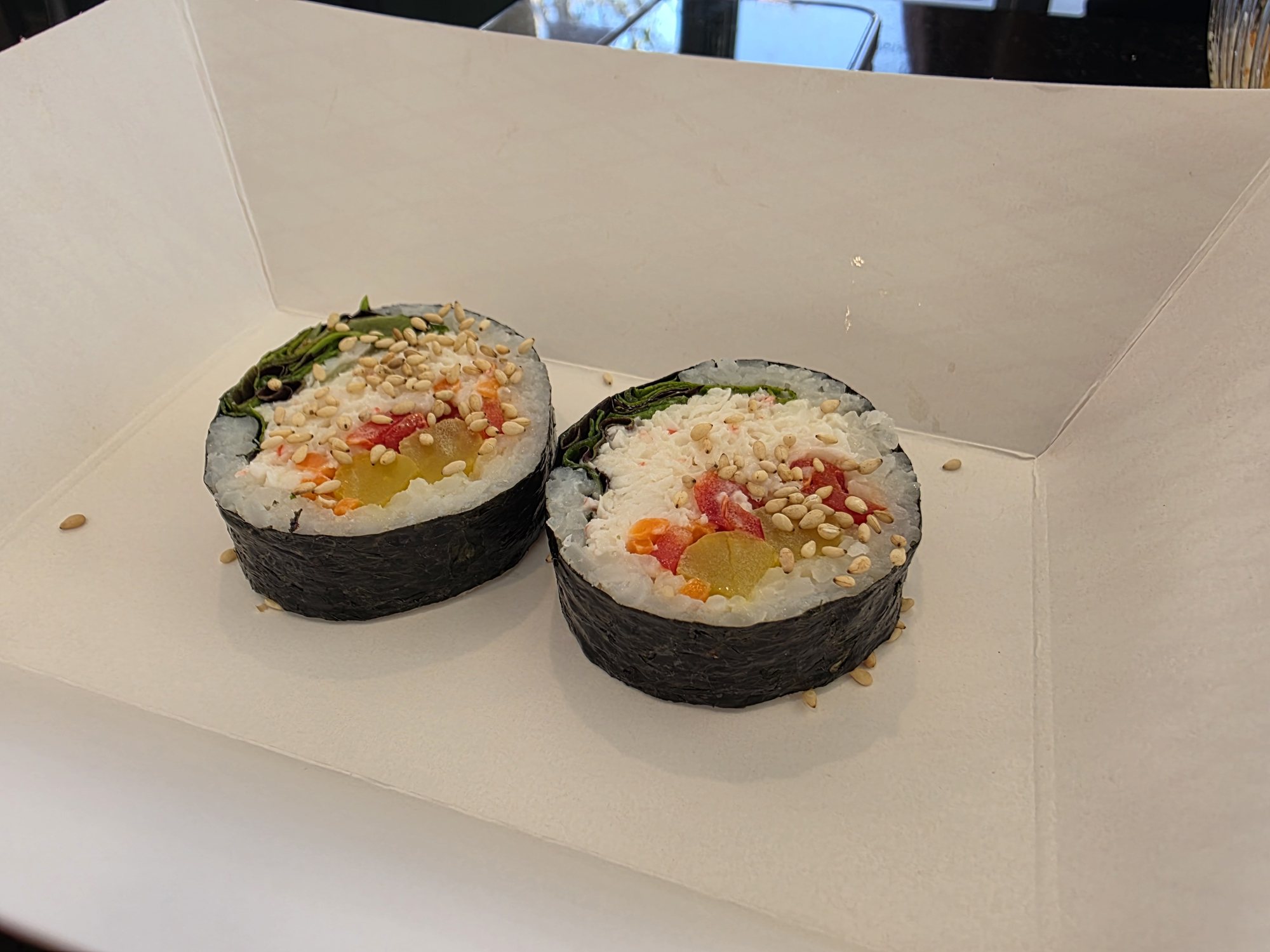 Two thick rounds of a K-roll, Korean-style kimbap wrapped in seaweed with colorful fillings and a crunchy topping, in a takeout box