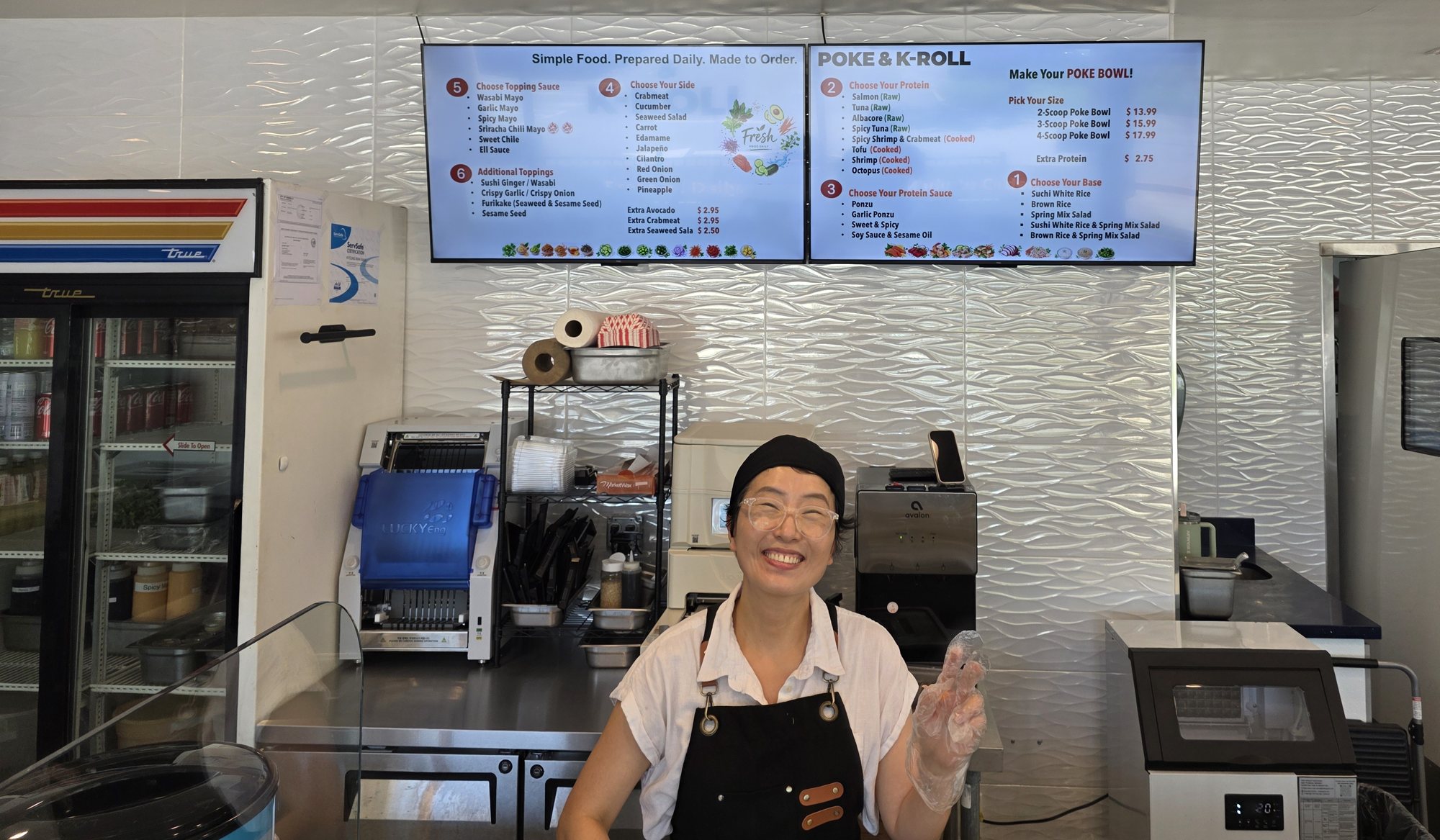 The owner of Poke & K-Roll smiling and waving from behind the counter, under the menu boards