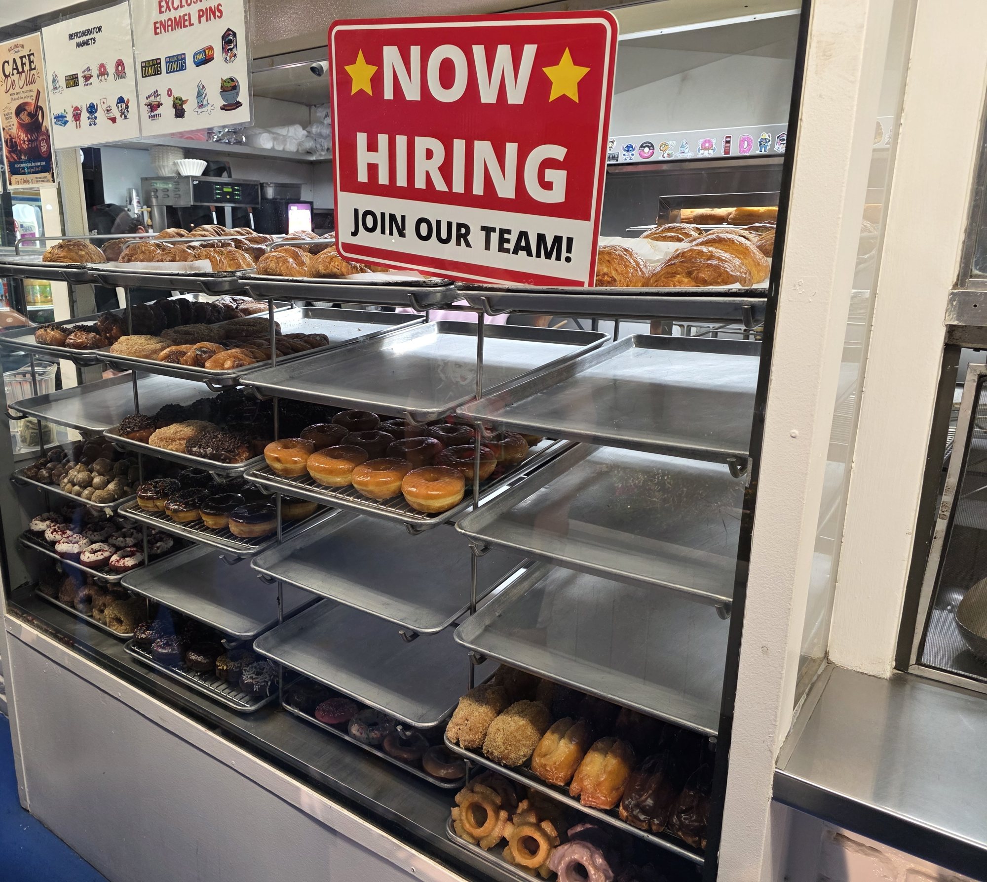 A red Now Hiring, Join Our Team sign on top of a donut case filled with trays of croissants and donuts at Rolling Pin Donuts in Camarillo